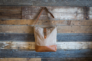 Beige canvas satchel with a leather bottom, hanging by its shoulder strap